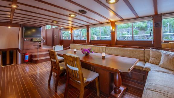 A cosy dining area inside a sailing boat with wooden furniture and large windows.